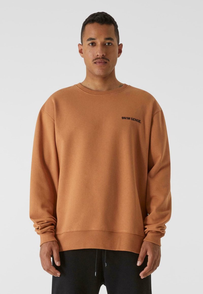 Felpa marrone realizzata in tessuto morbido, con scollo rotondo e maniche lunghe. Testo del logo nero sul petto sinistro. Vestibilità rilassata.