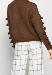 Pull en tricot marron avec des accents de pompons le long des manches, associé à un pantalon à carreaux blancs avec des lignes bleues.