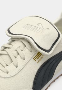 Hvide ruskinds sneakers med en sort stribe, der har en polstret tunge og guld logo-detalje. Snørerne er glatte og tæt vævet.
