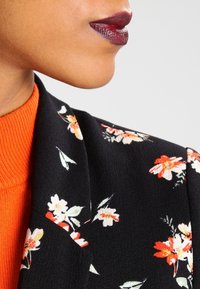 Svart blommig kavaj med mönster i vitt, orange och grönt, parat med en orange ribbad tröja under. Mörkläppstift accentuerar looken.