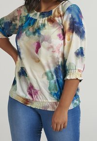 Blouse multicolore avec un motif aquarelle, encolure et bords des manches élastiqués, confectionnée en tissu léger. Associée à un jean bleu.