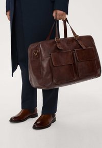Ruskea nahkainen duffel-laukku, jossa on kaksi etutaskua, ylätangot ja solkiyksityiskohtia. Sujuva pinta, suorakulmainen muoto ja minimaalinen brändäys.