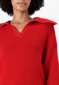 Red knitted sweater with a V-neck and oversized collar. Features a small logo on the chest and textured fabric with a loose fit.