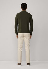 Polo a maniche lunghe di colore verde oliva con colletto, polsini con bottone, abbinato a pantaloni beige e scarpe marroni, visto da dietro.