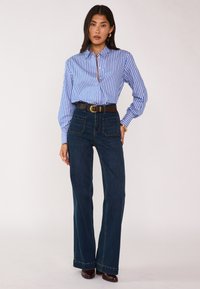 Blusa a righe blu e bianche con colletto, abbinata a jeans a zampa d'elefante in denim scuro con tasche frontali, una cintura marrone e scarpe marroni.