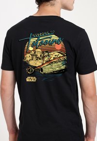 Černé tričko s retro grafikou s nápisem "Vítejte na Tatooine", speederem a pouštní krajinou. Výrazné barvy a stylizovaný design.
