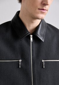 Melna jaka ar ādas apkakli, sudraba rāvējslēdzējiem un diviem priekšējiem kabatām ar rāvējslēdzēju aizvēršanu, izgatavota no teksturēta auduma.