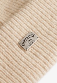 Kremowa dzianina prążkowana z metalową etykietą w kolorze srebrnym z napisem "SUPERDRY NYC Athletic". Skup się na fakturze i szczegółach szycia.