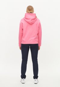 Růžová mikina s velkou kapucí, měkkou texturou a žebrovanými manžetami, v kombinaci s tmavomodrými kalhotami a bílými teniskami. Pohled zezadu.