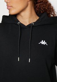 Primo piano di una persona che indossa un hoodie nero con un logo bianco di due figure sedute schiena contro schiena sul petto e cordini neri con punte metalliche.