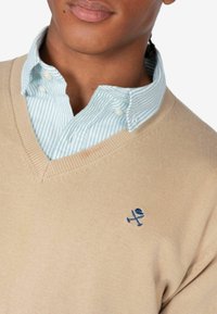 Beiger V-Ausschnitt-Pullover über einem hellblauen gestreiften Hemd mit Kragen. Besticktes Logo in Marineblau auf der linken Seite des Pullovers. Weiche Textur.