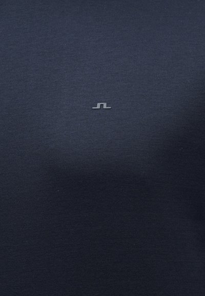 Σκούρο ναυτικό μπλε ύφασμα με ένα μικρό, κεντρικά τοποθετημένο ανοιχτό γκρι αφηρημένο λογότυπο ραμμένο κοντά στην επάνω μέση περιοχή.