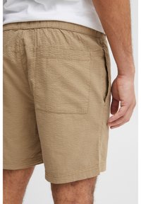 Khaki shorts with a textured, waffle-like pattern. Features an elastic waistband and side pockets, shown from the back with a hand resting on the fabric.