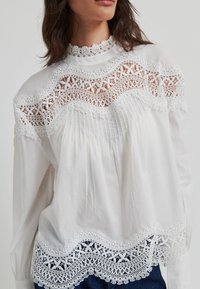 Blouse blanche avec bordure en dentelle délicate le long de l'encolure et de l'ourlet, avec des détails froncés et des manches longues pour une coupe ample.