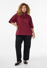 Lila Zip-Up-Oberteil mit kurzen Ärmeln, kombiniert mit schwarzen weit geschnittenen Hosen und schwarzen flachen Schuhen. Glattes Material mit einer entspannten Passform.