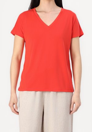 Kobieta w jasnoczerwonej koszulce z krótkim rękawem i dekoltem w serek oraz luźnych, jasnobeżowych spodniach, stojąca na białym tle.