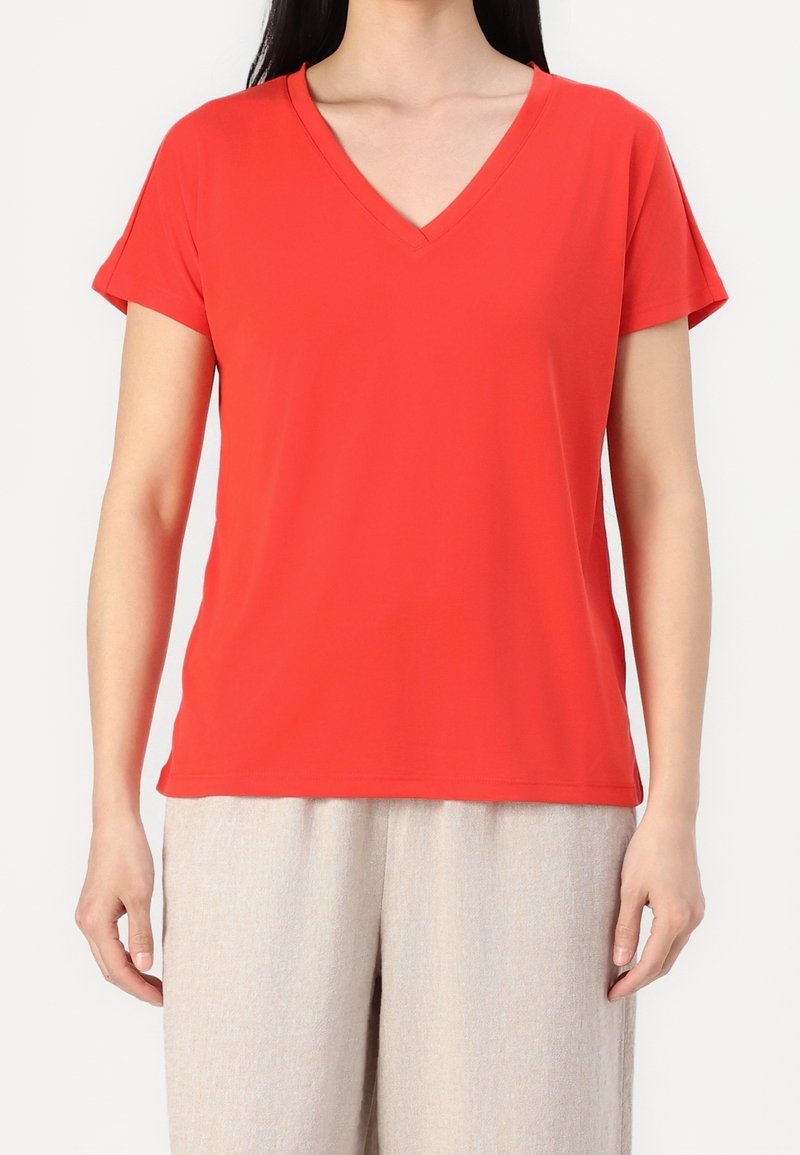 Femme portant un t-shirt rouge vif à col en V et manches courtes, avec un pantalon ample beige clair, debout devant un fond blanc.