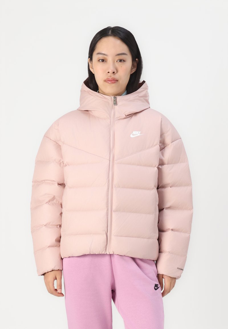 Puffige rosa Jacke mit Kapuze, mit gesteppten Abschnitten und einem Reißverschluss vorne. Weißes Logo auf der Brust. Kombiniert mit passenden rosa Hosen.