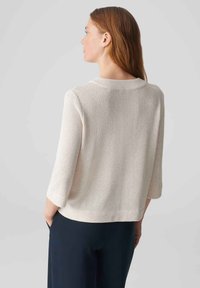Helle beige Strickoberteil mit dreiviertel Ärmeln und rundem Halsausschnitt. Strukturierten Stoff, lockere Passform und seitliche Schlitze. Kombiniert mit dunklen Hosen.