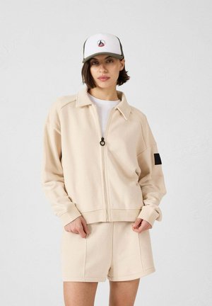 Giacca beige con zip e colletto, con un accento nero sulla manica, abbinata a pantaloni corti beige. Il modello indossa un cappello bianco con visiera verde.