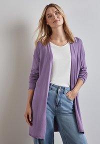 Cardigan violet clair en matière douce, avec un devant ouvert long, des poignets côtelés, porté sur un haut blanc et un jean bleu taille haute.
