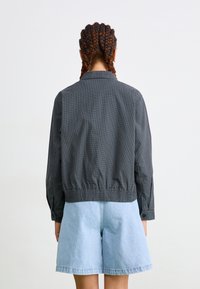 Svart rutete bomberjakke med ribbkant, kombinert med lyseblå denimshorts. Stoffet har en glatt tekstur og en avslappet passform.