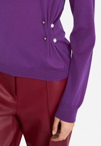 Maglione viola a maglia con arricciature laterali e dettagli decorativi in perle; abbinato a pantaloni rossi in ecopelle. Texture liscia e design aderente.