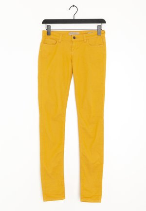 Gelbe Slim-Fit-Hose an einem schwarzen Bügel hängend, mit Vorderentaschen und einer Knopfleiste.