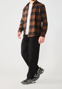 Camisa de franela a cuadros marrón y negro sobre una camiseta blanca, combinada con pantalones negros y zapatillas deportivas grises con acentos verdes.