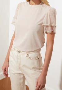 Top beige chiaro con maniche corte a sbuffo realizzato in tessuto trasparente, con bordo di pizzo. Abbinato a jeans beige chiaro a vita alta.