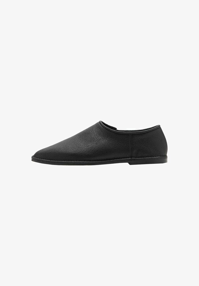 Zapato de piel negra tipo mocasín con punta redondeada y diseño minimalista. Presenta una textura suave y suela de goma de perfil bajo.