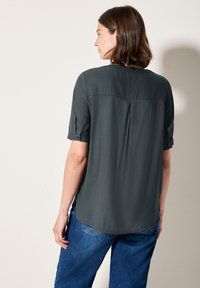 Blusa corta maniche, grigia scura e dal taglio morbido, con una piega sul retro come dettaglio di design, abbinata a jeans blu a gamba larga.