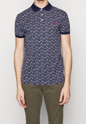 Granatowa koszulka polo z drobnym białym kwiatowym wzorem, żebrowanym kołnierzykiem i listwą z trzema guzikami. Na piersi małe czerwone logo.