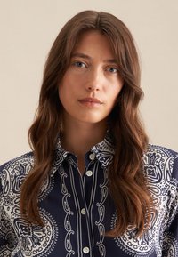Camicia button-up blu navy con motivo paisley bianco e disegni intricati, maniche lunghe e colletto classico. La texture del tessuto è morbida e visibile.