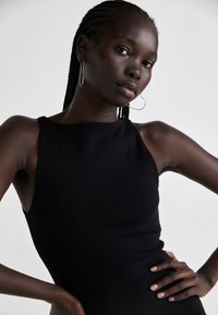 Rochie neagră fără mâneci din material ribbed, cu un guler înalt și o siluetă ajustată. Modelul poartă cercei mari tip cerc și are părul împletit.