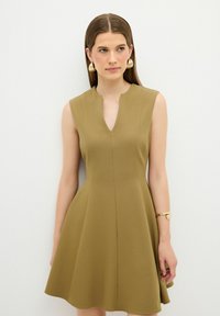 Ärmelloses A-Linien-Kleid aus olivgrünem Stoff mit tiefem V-Ausschnitt und weicher, glatter Textur. Akzentuiert mit goldenem Schmuck.