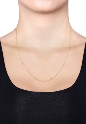 Collier délicat en chaîne d'or avec de petites perles rondes espacées régulièrement, porté autour du cou d'une femme au-dessus d'un haut noir à col rond plongeant.