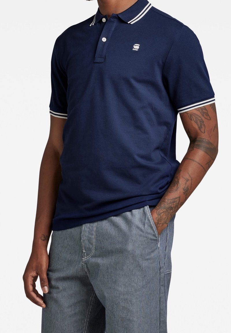 Homme portant un polo bleu marine avec bordure blanche au col et aux manches, pantalon gris rayé et tatouages sur les bras, debout la main dans la poche.