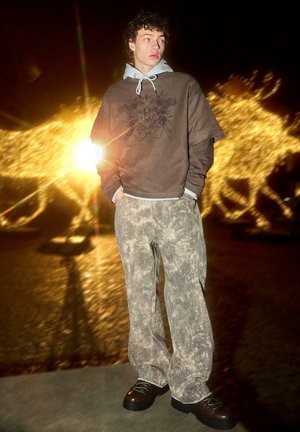 Felpa marrone indossata sopra una felpa con cappuccio grigio chiaro, abbinata a pantaloni beige fantasia e stivali scuri. Tessuti morbidi con una vestibilità comoda.