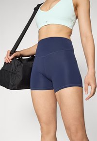 Navy high-waisted athletic shorts made of stretchy fabric. Features contrasting stitching and a smooth texture. Black gym bag held with one hand.