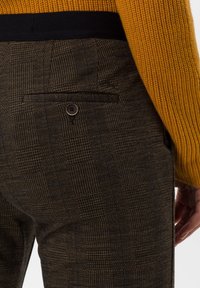 Pantalon marron à motifs avec un design à carreaux, une poche arrière avec un bouton, et une ceinture noire, assorti à un pull jaune texturé.