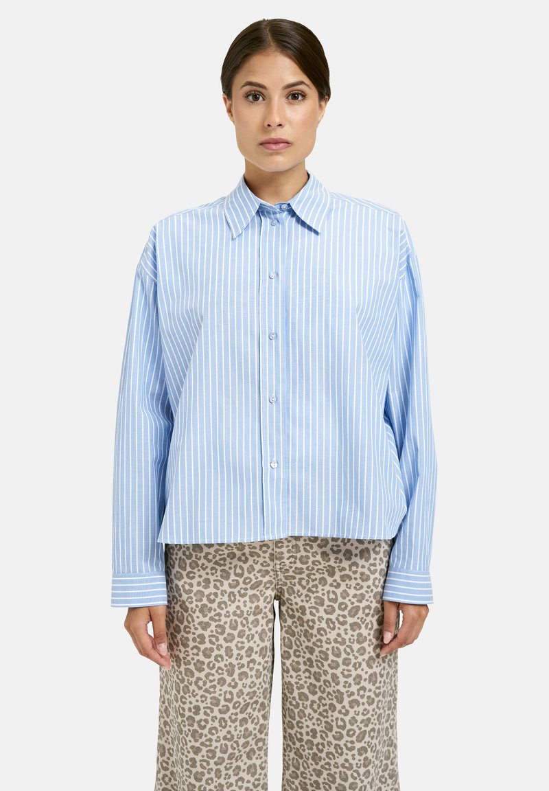 Woman standing facing forward wearing a blue and white vertical striped button-up shirt and beige leopard print pants.
