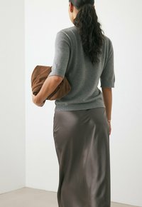 Maglione grigio a maniche corte abbinato a una gonna in satin lunga fino al pavimento di una tonalità scura, mentre tiene una pochette in tessuto marrone.
