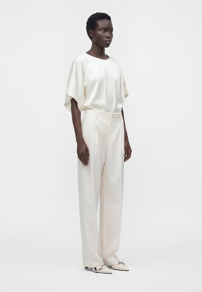 Femme aux cheveux courts portant un chemisier ample en satin blanc, un pantalon blanc taille haute à jambes larges et des chaussures blanches pointues avec une boucle.