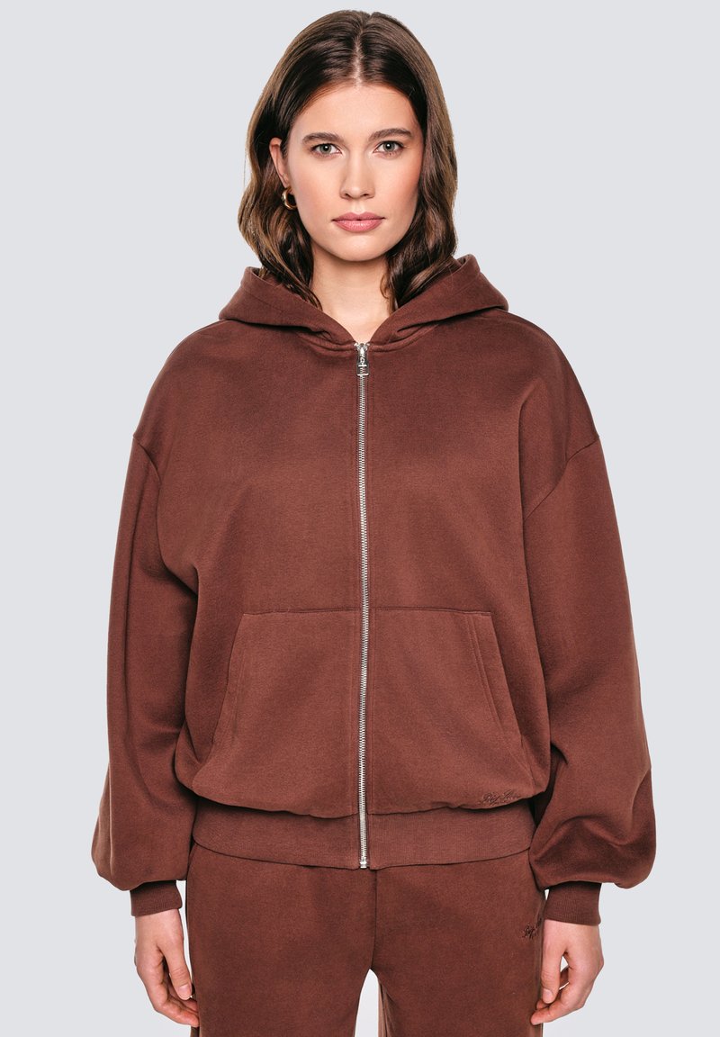 Pruun zip-up kapuutsiga dressipluus, mis on valmistatud pehmest kangast, sisaldab kapuutsi, esipokke ja soonikuga varrukaid. Lihtne disain, ilma nähtavate mustrite või aktsentideta.