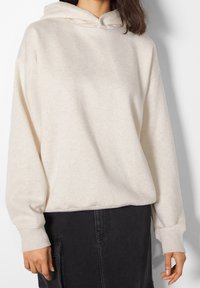 Krämfärgad hoodie med mjuk textur, avslappnad passform och kenguruficka, tillsammans med en svart kjol. Har en huva med dragsko.