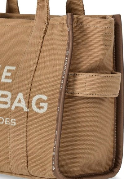 Kangaskassi, jossa on tan-värinen kangas, vahvistetut ruskeat reunat ja valkoisella painatuksella osittain lukee "BAG" sivutaskussa.