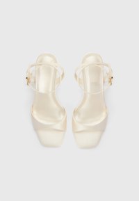 Beige satin hæle sandaler med to diagonale stropper, en ankelrem, firkantet tå og en glat tekstur. Guldfarvet hardware på spænde.