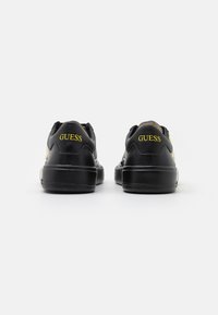 Black leather sneakers with a smooth texture, featuring a yellow "GUESS" logo on the heel and side, rounded toe, and rubber sole.