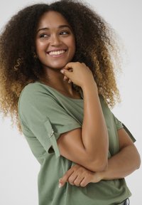 Green short-sleeve top with rolled cuffs and silver hardware accents. Soft fabric with a relaxed fit. Model poses with a smile.